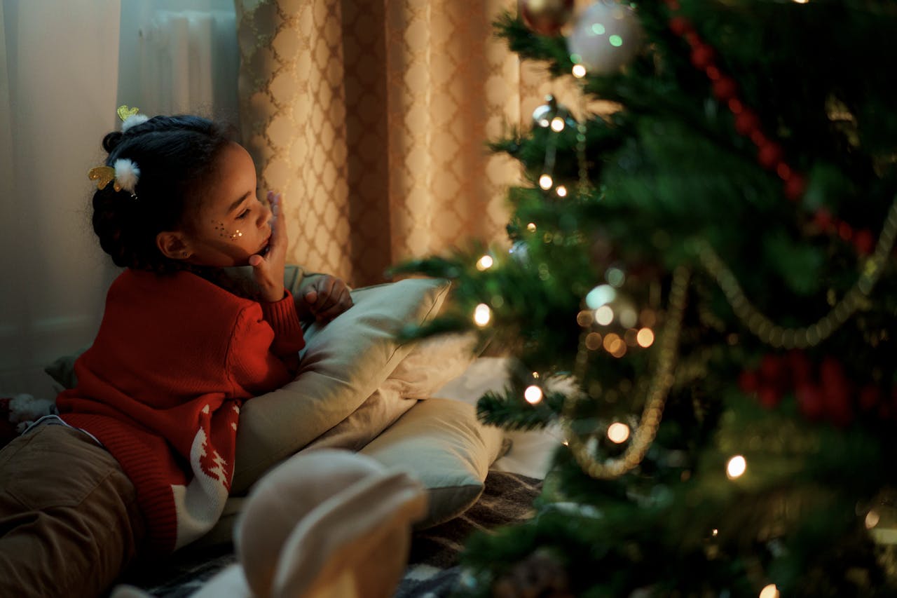 hero-img-01 Child lying by a decorated Christmas tree, gazing with wonder and holiday joy indoors.